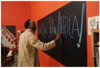 Still from the film The Inheritance. A Black man in a tan coat writes John Africa on a chalkboard.