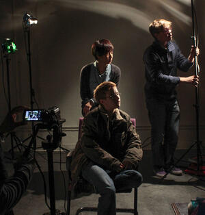 Image from Lighting Workshop, three people are on a film set, one sits in a chair, one is standing behind the seated person adjusting a light and another standing person adjusts a film production light on the right.